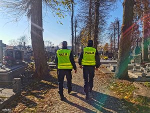 na zdjęciu dwóch umundurowanych policjantów w kamizelkach odblaskowych idących po cmentarzu