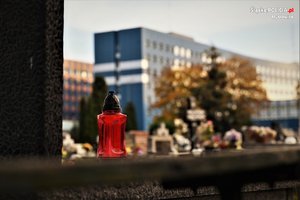 Na zdjęciu czerwony znicz, a w tle cmentarz i budynek Komendy Wojewódzkiej Policji w Katowicach
