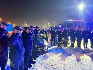 Policjanci wraz ze strażakami, stojąc przy swoich wozach z włączonymi sygnałami, oddają honory