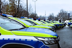 Zdjęcie. Rząd oznakowanych radiowozów zaparkowanych przed budynkiem Komendy Wojewódzkiej Policji w Katowicach, obok których stoją umundurowani policjanci.