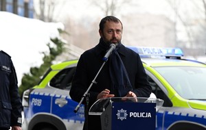 Zdjęcie. Mężczyzna przemawia przed mikrofonem przed budynkiem komendy, w tle radiowóz