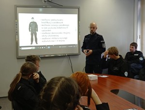 Zdjęcie. Grupa uczniów w sali, w której umundurowany policjant prowadzi wykład obok tablicy z wyświetlona prezentacją