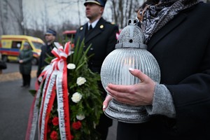 Na zdjęciu znicz trzymany w kobiecych dłoniach, obok strażak trzymający kwiaty.