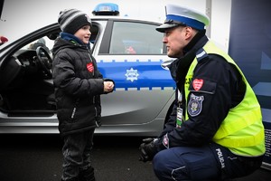 Zdjęcie. Policjant przy radiowozie kuca naprzeciwko chłopca.
