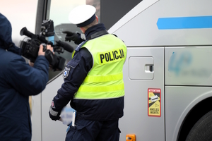 Zdjęcie przedstawia umundurowanego policjanta przy autokarze. W tle osoba z kamerą.