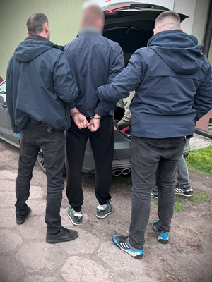 Na zdjęciu widać dwóch policjantów w ubraniu cywilnym prowadzących zatrzymanego mężczyznę w kajdankach.