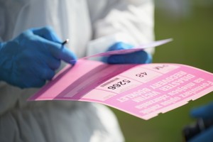 na zdjęciu fragment sylwetki osoby w białym kombinezonie i niebieskich rękawiczkach, w dłoniach różowy formularz i długopis