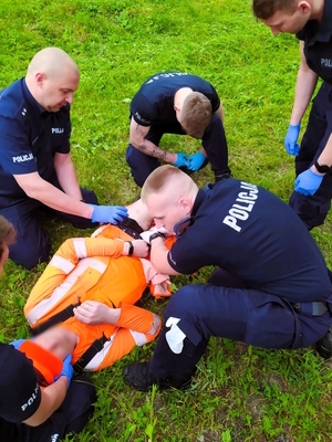 Na zdjęciu grupa policjantów udziela pierwszej pomocy poszkodowanemu, leżącemu mężczyźnie.