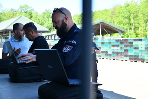 na zdjęciu policjant pracujący przy laptopie oraz dwóch mężczyzn w ubraniach cywilnych, z których jeden pracuje przy laptopie a drugi stoi.