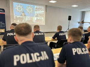 Na zdjęciu siedzący w ławkach uczestnicy szkolenia zawodowego podoficerskiego w trakcie wykładu z nauczycielem policyjnym. Przed uczestnikami wyświetlony jest na ekranie obraz z połączenia zdalnego ze Szkołą Policji w Katowicach.