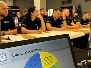 Zbliżenie na ekran komputera na którym wyświetlana jest prezentacja. W tle widać uczestników szkolenia zawodowego podoficerskiego.