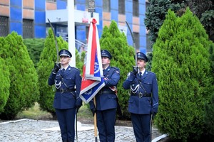 Policjanci w umundurowaniu galowym prezentują poczet sztandarowy na zewnątrz budynku