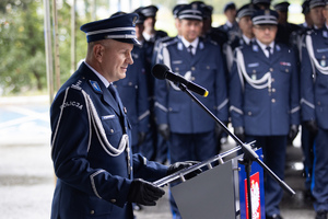 Policjanci w umundurowaniu galowym stoją w szeregu, a generał przemawia do mikrofonu