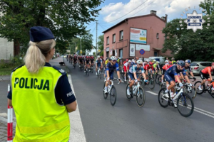 Zdjęcie kolorowe przedstawiające policjantów podczas wyścigu kolarskiego. Na zdjęciu z lewej strony stoi policjantka w odblaskowej kamizelce. Drogą jedzie peleton.