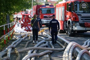 Na zdjęciu widoczny tyłem umundurowane policjant idący drogą wzdłuż rozłożonych węży strażackich