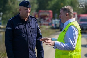 Na zdjęciu widoczny umundurowany policjant rozmawiający z drugim w kamizelce odblaskowej