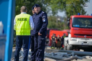 Na zdjęciu widoczni dwaj umundurowani policjanci rozmawiający z trzecim w kamizelce odblaskowej z napisem Policja