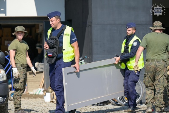 Na zdjęciu widoczni dwaj umundurowani policjanci przenoszący drewniany element - obok stoją żołnierze WOT