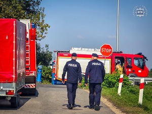 Na zdjęciu dwaj umundurowani policjanci, idący obok wozów strażackich