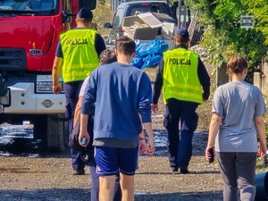 Na zdjęciu dwaj umundurowani policjanci w kamizelkach odblaskowych idący wśród ludzi obok wozów strażackich