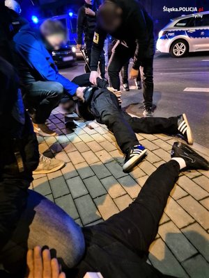 Na zdjęciu mężczyzna leżący na chodniku przytrzymywany przez nieumundurowanych policjantów. Obok nogi drugiego z zatrzymanych mężczyzn. W tle radiowozy policyjne.