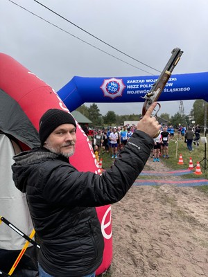 Na zdjęciu zbliżenie na przedstawiciela Wydziału Doboru i Szkolenia KWP w Katowicach, który trzyma w dłoni replikę Centaky Patriot przed oddaniem symbolicznego strzału w powietrze rozpoczynającego bieg. W tle widać uczestników przygotowanych do startu.