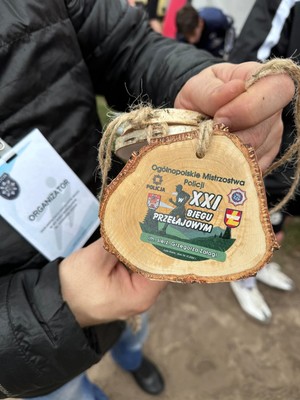 Zbliżenie na pamiątkowy medal  Ogólnopolskich Mistrzostw Policji w XXI Biegu Przełajowym im. Grzegorza Załogi trzymany w dłoniach przedstawiciela organizatora.