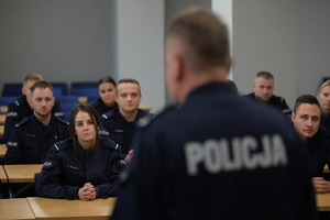 Zdjęcie przedstawia umundurowanych uczestników kursu podoficerskiego siedzących przy stolikach, słuchających przemawiającego do nich oficera Policji.