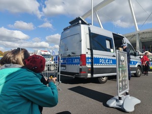 Zdjęcie przedstawiające policyjny radiowóz, a na pierwszym planie kobietę z dzieckiem Na