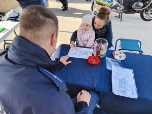 Na zdjęciu widać dziecko z mamą rysujące kolorowankę, tyłem policjant