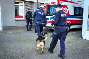 Zdjęcie przedstawia czterech policjantów a w tym jeden prowadzi policyjnego psa a w tle jest widoczna karetka