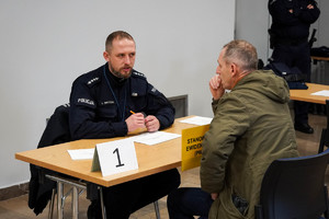 na zdjęciu mężczyzna siedzący przy stole, na przeciwko siedzący policjant, mężczyźni są w trakcie rozmowy