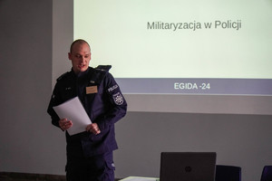 na zdjęciu policjant w trakcie wypowiedzi, za nim wyświetlony slajd z napisem Militaryzacja w Policji EGIDA 24