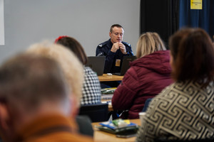 na zdjęciu policjant w trakcie wypowiedzi siedzący przy stoliku przed zgromadzonymi osobami