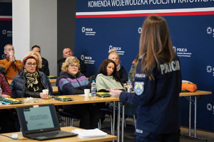 na zdjęciu policjantka stojąca przed zgromadzonymi osobami siedzącymi przy stołach