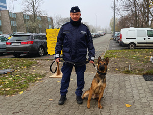 na zdjęciu policjant i pies brązowego koloru, policjant trzymający psa na smyczy, za nimi tabliczka informacyjna dotycząca EGIDY 24, parking samochodowy