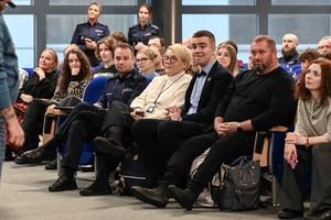 na zdjęciu siedzące na krzesłach osoby i policjanci.