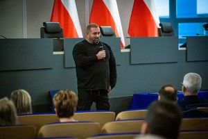 na zdjęciu mężczyzna przemawiający do mikrofonu przed zgromadzonymi na sali konferencyjnej osobami.