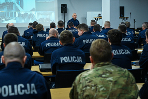 Zdjęcie przedstawia: słuchaczy siedzących w ławkach. Przed nimi stoi umundurowany policjant i przemawia.