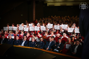 osoby siedzące na widowni trzymają kartki z imionami osób i ich wiekiem, które zginęły na drodze