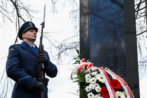 Na zdjęciu policjant w mundurze galowym na tle Pomnika upamiętniającego zmarłych w katastrofie MTK