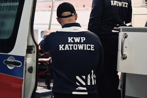 na zdjęciu widoczni są dwaj umundurowani policjanci stojący tyłem do zdjęcia przed otwarta karetką na kurtce maja napis KWP Katowice