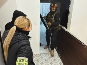 na zdjęciu policyjny kontrterrorysta z taranem w rękach w drzwiach mieszkania, obok po cywilu funkcjonariusze straży granicznej