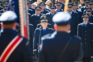 na pierwszym planie mniej wyraźni policjanci stojący tyłem przed nimi wyraźnie widoczny komendant wojewódzki policji w Katowicach podczas przemówienia w tle za nim zaproszeni goście