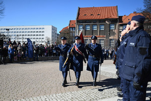 zdjęcie obejmujące większość placu po prawej stronie stoją policjanci mający ślubowanie środkiem idą policjanci z pocztem sztandarowym w tle zaproszeni goście za nimi budynki