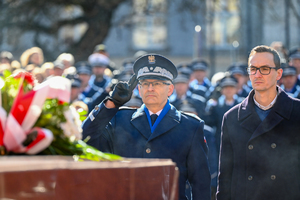 na pierwszym planie fragment wiązanki w tle komendant wojewódzki oddający honor przed pomnikiem