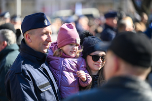 zbliżenie na policjanta obok którego stoi kobieta która trzyma na rękach dziecko