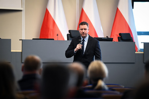 Zdjęcie przedstawia mężczyznę w garniturze przemawiającego do publiczności.