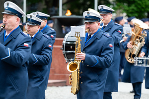 Grafika przedstawia umundurowaną orkiestrę Policji na Placu Chrobrego w Katowicach.