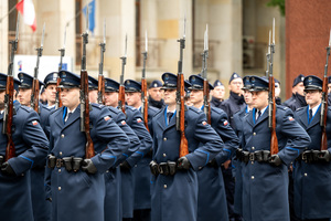 Grafika przedstawia policjantów kompanii honorowej w płaszczach galowych , trzymających w rękach bagnety.
